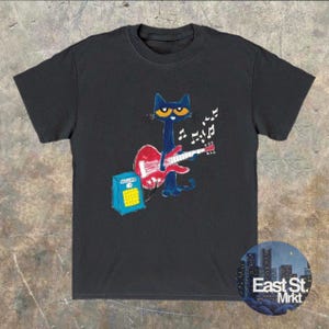 May include: Dark grey t-shirt with a cartoon cat playing a red guitar, accompanied by musical notes. A blue amplifier is positioned nearby. The shirt includes a round logo that reads "East St. Mrkt".