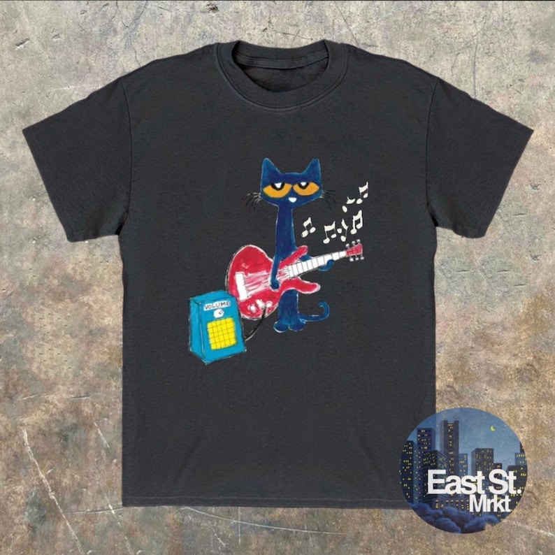 May include: Dark grey t-shirt with a cartoon cat playing a red guitar, accompanied by musical notes. A blue amplifier is positioned nearby. The shirt includes a round logo that reads "East St. Mrkt".