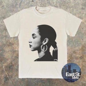 May include: Cream-colored t-shirt featuring a black and white portrait of a woman with hoop earrings. The design is a side profile of the woman's face. A circular logo with the text "East St. Mrkt" is on the bottom right.