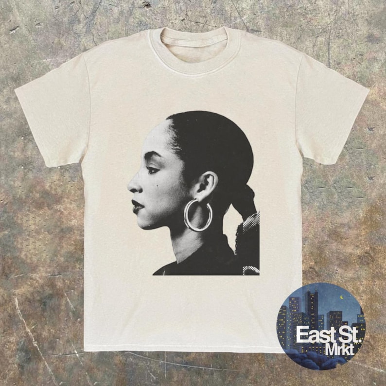 May include: Cream-colored t-shirt featuring a black and white portrait of a woman with hoop earrings. The design is a side profile of the woman's face. A circular logo with the text "East St. Mrkt" is on the bottom right.