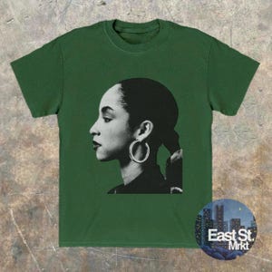 May include: Green t-shirt featuring a black and white profile portrait of a woman wearing hoop earrings. The image is centered on the shirt, with the text "East St. Mrkt" in a circular design at the bottom right.