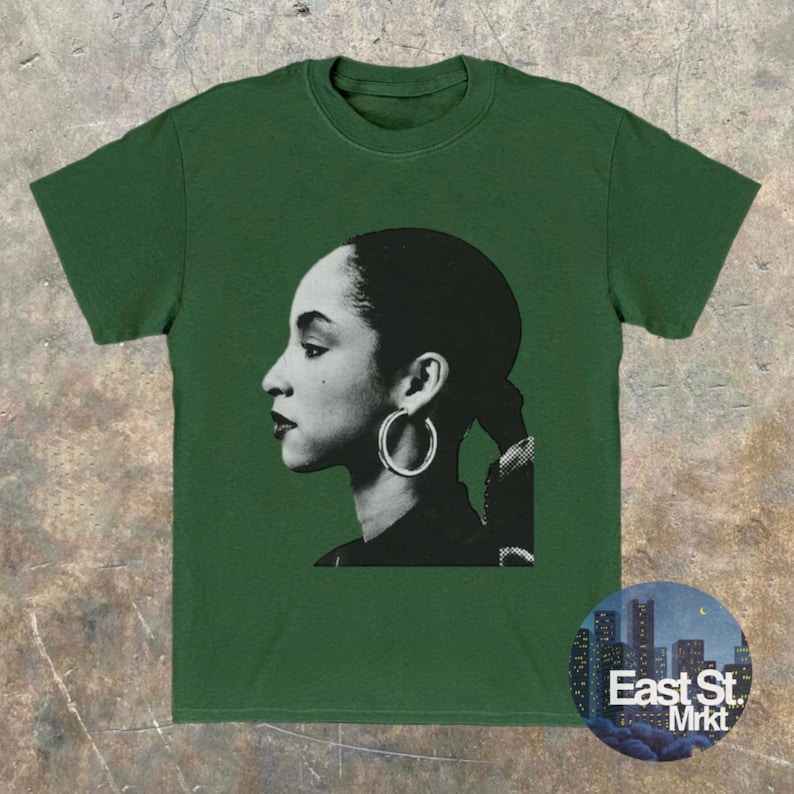 May include: Green t-shirt featuring a black and white profile portrait of a woman wearing hoop earrings. The image is centered on the shirt, with the text "East St. Mrkt" in a circular design at the bottom right.
