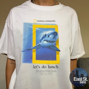 Op de afbeelding: Een wit t-shirt met een National Geographic-afbeelding van een witte haai die uit een geel rechthoekig kader komt. Onder de haai staat de tekst: let's do lunch. The Great White Shark, Weight: up to 1.179 kg, Length: up to 5,9 meter.