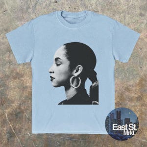 May include: Light blue t-shirt featuring a black and white portrait of a woman with hoop earrings. The image is a side profile. A circular logo with the text "East St. Mrkt" is on the bottom right.