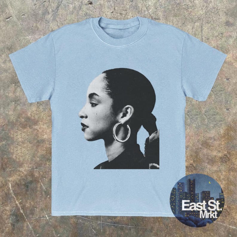May include: Light blue t-shirt featuring a black and white portrait of a woman with hoop earrings. The image is a side profile. A circular logo with the text "East St. Mrkt" is on the bottom right.