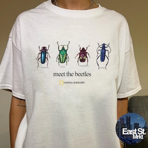 Op de afbeelding: Een wit T-shirt met korte mouwen en een afbeelding van vier kleurrijke kevers op een rij. Onder de insecten staat de tekst "meet the beetles" in het zwart, gevolgd door het National Geographic logo in geel en zwart.