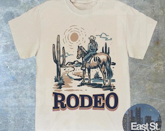 May include: A beige t-shirt featuring a graphic print of a woman riding a horse in a desert landscape. The design includes cacti, a sun, and the word "RODEO" in blue lettering.