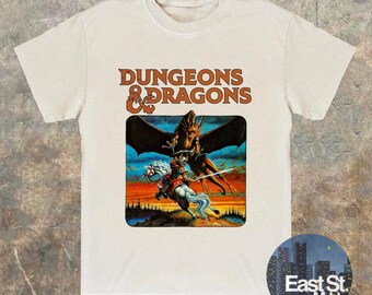 May include: Beige t-shirt featuring the text "DUNGEONS & DRAGONS" in brown. The shirt has a colorful graphic of a knight on a white horse fighting a dragon. The shirt is a crew neck and has short sleeves.
