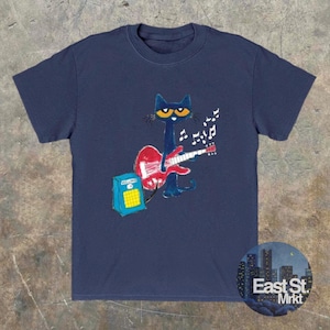May include: Navy blue t-shirt with a cartoon cat playing a red guitar, accompanied by musical notes. The cat has yellow eyes and is positioned in front of a blue amplifier. The shirt features a round logo that says "East St. Mrkt".