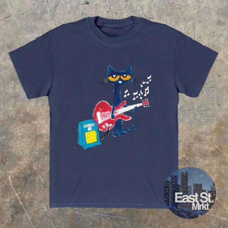 May include: Navy blue t-shirt with a cartoon cat playing a red guitar, accompanied by musical notes. The cat has yellow eyes and is positioned in front of a blue amplifier. The shirt features a round logo that says "East St. Mrkt".