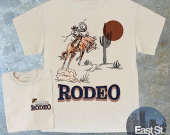 May include: A beige t-shirt featuring a graphic of a cowboy riding a bucking horse. The text "RODEO" is printed in blue on the front of the shirt. The back of the shirt has the text "Cowboy Rodeo" printed in brown.