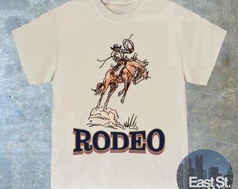 May include: A beige t-shirt featuring a graphic of a cowboy riding a horse. The graphic is in brown and black. The text "RODEO" is printed in blue with a gold outline.