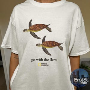 Może przedstawiać: Biały t-shirt z krótkim rękawem z grafiką przedstawiającą dwa pływające żółwie morskie. Poniżej żółwi znajduje się napis "go with the flow" czcionką szeryfową, a pod nim żółto-czarne logo National Geographic umieszczone na środku klatki piersiowej.