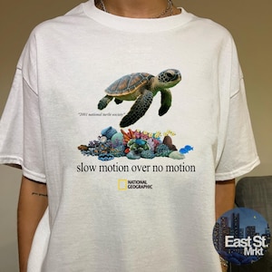 Puede incluir: Camiseta blanca con un gráfico de tortuga marina sobre corales coloridos y el texto "slow motion over no motion". El logotipo de National Geographic está debajo. La camiseta también tiene el texto "2001 national turtle society."