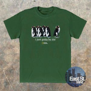 Może przedstawiać: Leśno-zielony t-shirt z kr&oacute;tkim rękawem z grafiką przedstawiającą pięć pingwin&oacute;w w rzędzie. Pod pingwinami znajduje się napis "i just gotta be me" oraz logo National Geographic. Koszulka posiada okrągły dekolt.