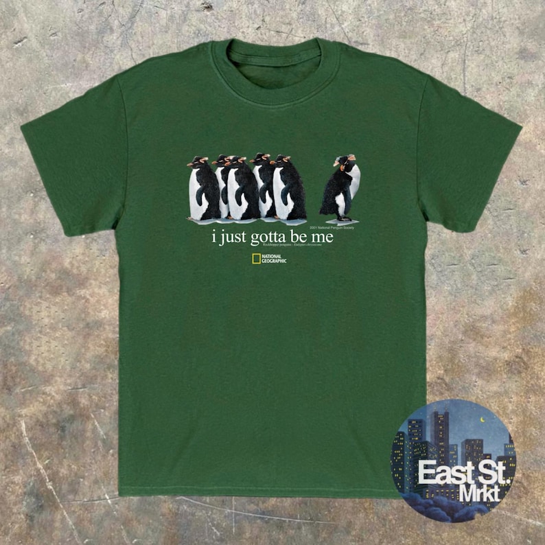 Może przedstawiać: Leśno-zielony t-shirt z kr&oacute;tkim rękawem z grafiką przedstawiającą pięć pingwin&oacute;w w rzędzie. Pod pingwinami znajduje się napis "i just gotta be me" oraz logo National Geographic. Koszulka posiada okrągły dekolt.