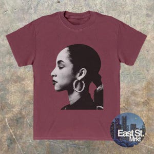 May include: Burgundy t-shirt featuring a black and white profile portrait of a woman wearing hoop earrings. The shirt has a crew neck and short sleeves. A circular logo with the text "East St. Mrkt" is on the bottom right.