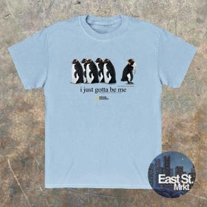 Może przedstawiać: Jasnoniebieski t-shirt z kr&oacute;tkim rękawem z grafiką przedstawiającą pięć pingwin&oacute;w stojących w rzędzie. Pod pingwinami nadrukowany jest napis "i just gotta be me" oraz logo National Geographic wraz z dodatkowym małym tekstem.