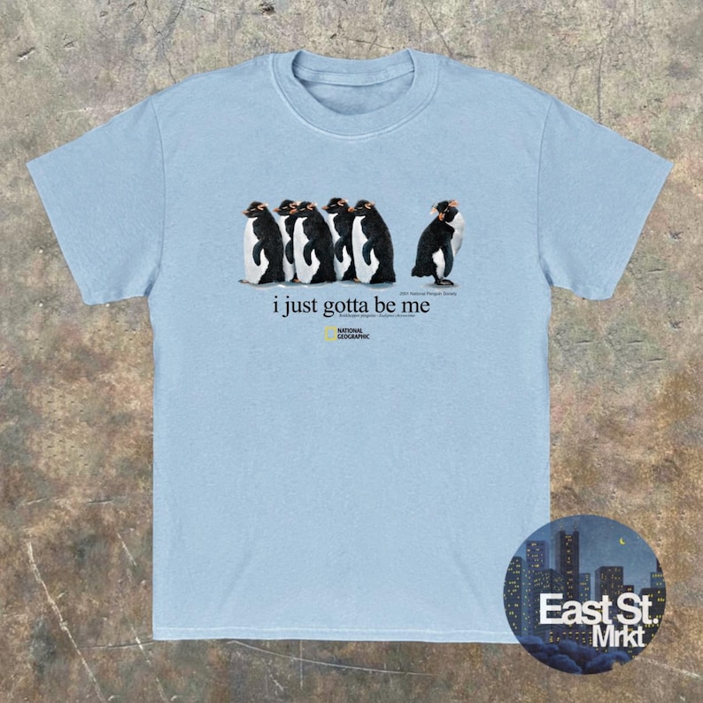 Może przedstawiać: Jasnoniebieski t-shirt z kr&oacute;tkim rękawem z grafiką przedstawiającą pięć pingwin&oacute;w stojących w rzędzie. Pod pingwinami nadrukowany jest napis "i just gotta be me" oraz logo National Geographic wraz z dodatkowym małym tekstem.