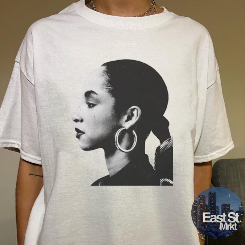 May include: White t-shirt featuring a black and white portrait of a woman with large hoop earrings. The image is a side profile of the woman's face and hair. The shirt has short sleeves and a round neckline.