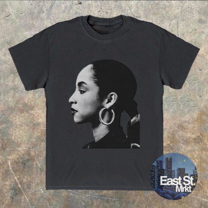 May include: Dark gray t-shirt featuring a black and white portrait of a woman with hoop earrings. The image is a side profile. The t-shirt has a round neckline and short sleeves. A circular logo with the text "East St. Mrkt" is on the bottom right.