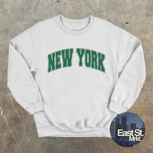 以下が含まれることがあります： 前面に緑色の「NEW YORK」のグラフィックがプリントされた白いスウェットシャツです。