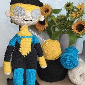 Könnte beinhalten: Handgefertigte Häkelpuppe eines Superhelden, mit schwarzem Barett, gelbem Haar und einem blau-schwarzen Anzug. Die Puppe hat graue Augenabdeckungen und blaue Stiefel. Sonnenblumen und Garn sind im Hintergrund.