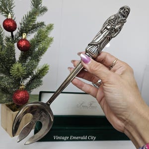 Könnte beinhalten: Silberfarbene Serviergabel mit einer detaillierten Weihnachtsmannfigur am Griff. Die Gabel hat einen langen, schlanken Griff und einen breiten, geschlitzten Kopf. Der Griff weist aufwendige Schnitzereien und ein dekoratives Design auf. Der Text "Vintage Emerald City" befindet sich unten.