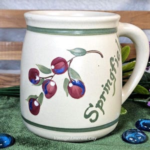 May include: Cream-colored ceramic mug with a green handle and rim. The mug features a painted branch of cherries in shades of red and blue, with green leaves. The word "Springfield" is written in green script on the side. "Vintage Emerald City" is written at the bottom.