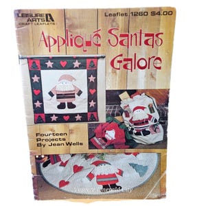May include: A Leisure Arts craft leaflet titled Appliqué Santas Galore, featuring fourteen projects by Jean Wells. The cover displays various Santa-themed fabric appliqué designs, including a wall hanging, a gift sack, and a tree skirt.