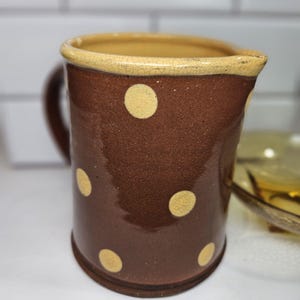 May include: A brown ceramic pitcher with a handle and a spout. The pitcher is decorated with large, evenly spaced yellow polka dots. The rim is a lighter tan color with a cracked texture.