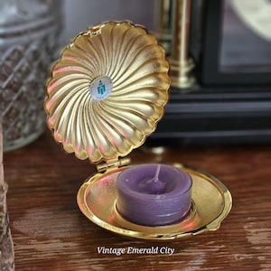 May include: A gold-colored, clam-shaped trinket box with a purple candle inside. The box is open, revealing the candle. The box has a fluted design and a small, round sticker on the top. The text "Vintage Emerald City" is at the bottom.