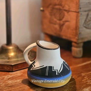 May include: A small ceramic pitcher with a white upper body and a black geometric pattern. The lower section features horizontal bands of blue, grey, and yellow. The handle is white, and the rim is outlined in black. The text "Vintage.Emerald City" is visible.