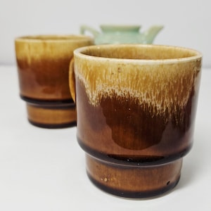 May include: Two brown ceramic mugs with a layered design. The mugs have a handle and a light brown rim that fades into a darker brown. A light green teapot is visible in the background.