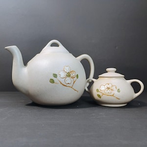 May include: A cream-colored ceramic teapot and sugar bowl set. The teapot has a curved spout and handle, and the sugar bowl has a lid. Both pieces are decorated with a floral design in brown and green.