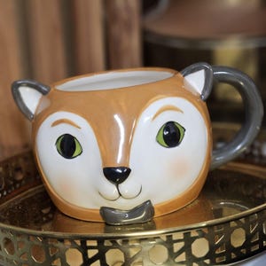 Op de afbeelding: Een keramische mok in de vorm van een vossengezicht. De mok heeft voornamelijk bruine en witte kleuren, met een grijze oor en oren. De vos heeft groene ogen en een zwarte neus. De mok staat op een goudkleurig dienblad.