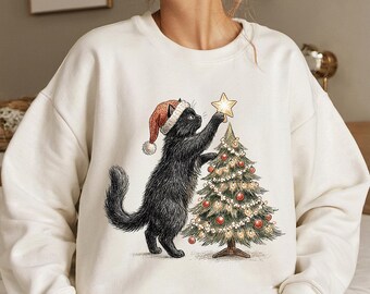 Puede incluir: Sudadera de color crema con un gato negro con un gorro de Papá Noel, alcanzando una estrella en la cima de un árbol de Navidad. El árbol está decorado con luces y adornos. Un diseño festivo navideño.