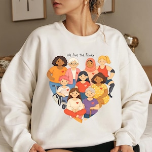 Può includere: Felpa bianca con un'illustrazione a forma di cuore di donne diverse. L'illustrazione include donne di diverse etnie, età e abilità. Il testo "We Are the Power" è stampato sopra l'illustrazione in nero.