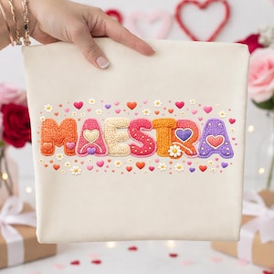 May include: A cream-colored tote bag with the word "MAESTRA" embroidered in colorful, textured letters. The letters are decorated with hearts and small flowers. The bag is held by a person.