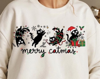 Puede incluir: Sudadera color crema con un diseño lúdico de "merry catmas". El gráfico incluye gatos negros decorados con luces navideñas, un gorro de Papá Noel y regalos. Copos de nieve y estrellas festivas completan el tema navideño.