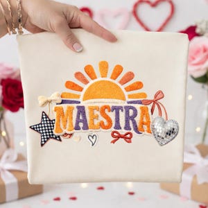 Può includere: Tessuto color crema con la parola ricamata "MAESTRA" in viola e arancione, sormontato da un disegno di sole. Gli accenti includono una stella in bianco e nero, un cuore e un cuore a sfera stroboscopica.