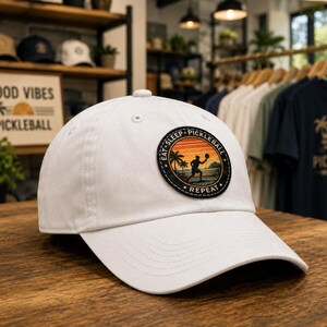 May include: A white baseball cap featuring a circular patch on the front. The patch displays a silhouette of a person playing pickleball against a sunset background with palm trees, surrounded by the text EAT SLEEP PICKLEBALL REPEAT.