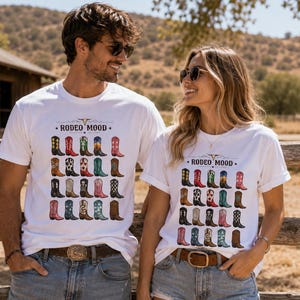 May include: A white short-sleeve t-shirt featuring a graphic print of twenty-five colorful cowboy boots arranged in a grid. Above the boots, the text reads RODEO MOOD with a small bull skull icon. The design includes various patterns and colors.