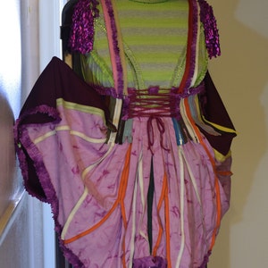 May include: A purple and pink dress with a green hat and a flower on top. The dress is made of different fabrics and has a lot of layers. It is a unique and creative design.