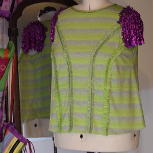 May include: A green and grey striped shirt with ruffled trim and purple fabric flowers on the shoulders.