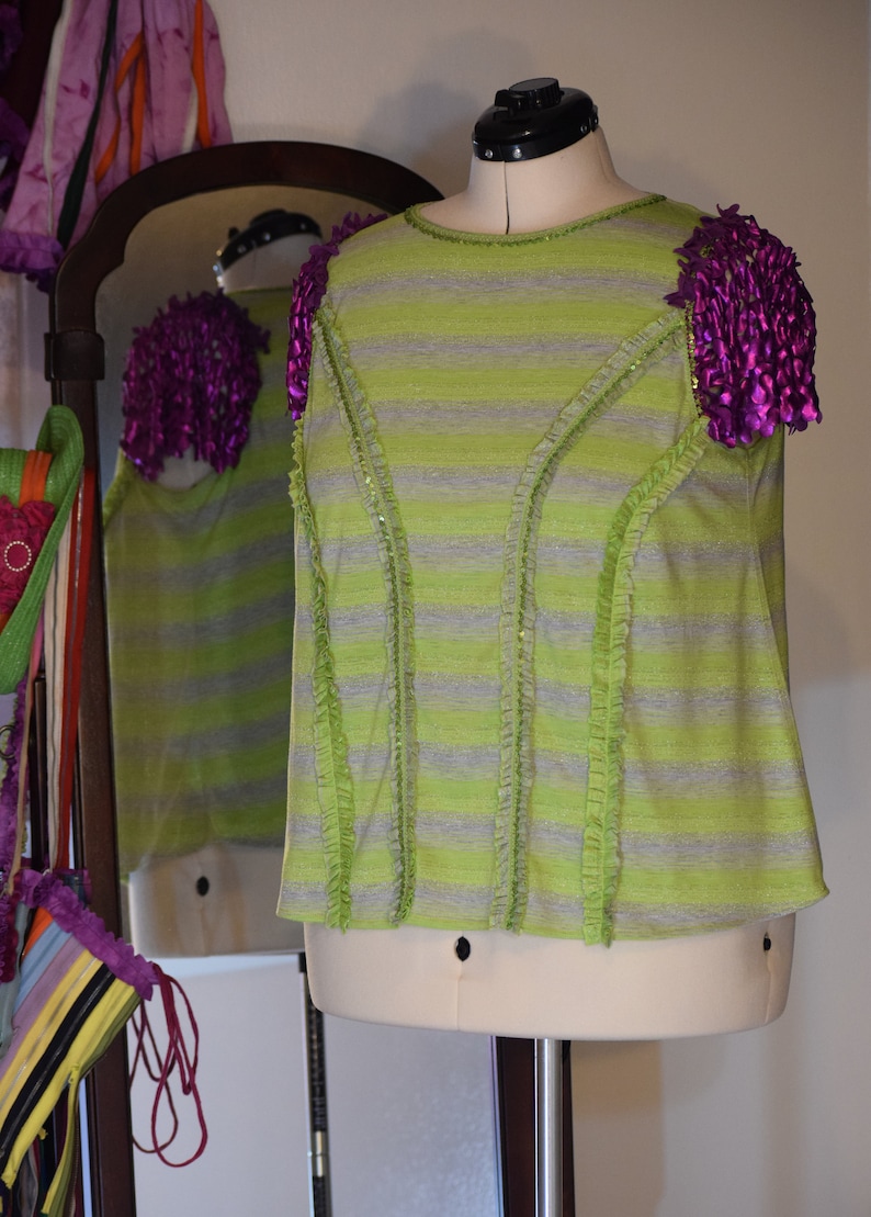 May include: A green and grey striped shirt with ruffled trim and purple fabric flowers on the shoulders.