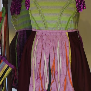 May include: A green and purple striped dress with a pink and orange layered skirt. The skirt is made of strips of fabric with a ruffled hem. The dress has purple fabric flowers on the shoulders.
