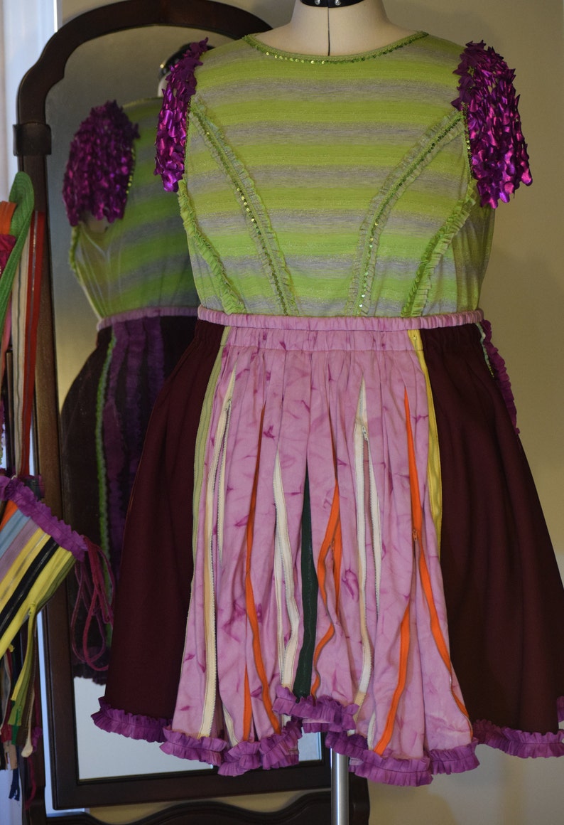 May include: A green and purple striped dress with a pink and orange layered skirt. The skirt is made of strips of fabric with a ruffled hem. The dress has purple fabric flowers on the shoulders.