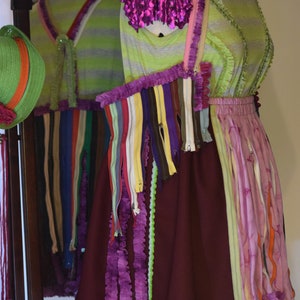May include: A purple, green, and brown dress with a ruffled hem and a variety of colorful zippers and fabric strips. The dress is made from recycled materials and has a unique, handmade aesthetic.