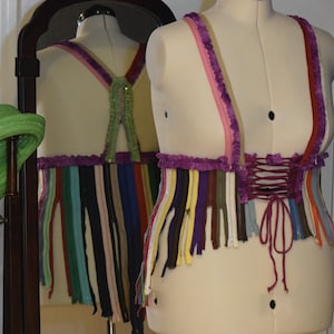 May include: A purple and white corset with a pink and white trim. The corset is made of a variety of colorful strips of fabric, possibly zippers. The corset is tied at the back with a burgundy ribbon.